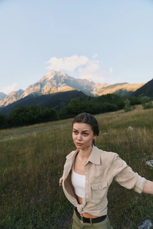Cheerful woman outdoors on a mountainside, wearing a casual jacket over a white top, in a sunlit meadow with natural colors and a light breeze, relaxed and confident