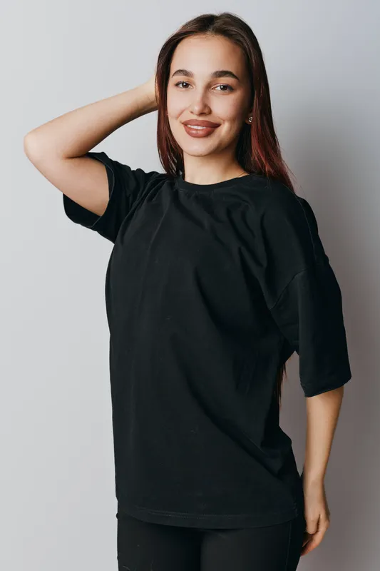Young woman with long hair wearing black t shirt standing and smiling with hand behind head on light background. Casual style, confident pose and natural look for portrait.
