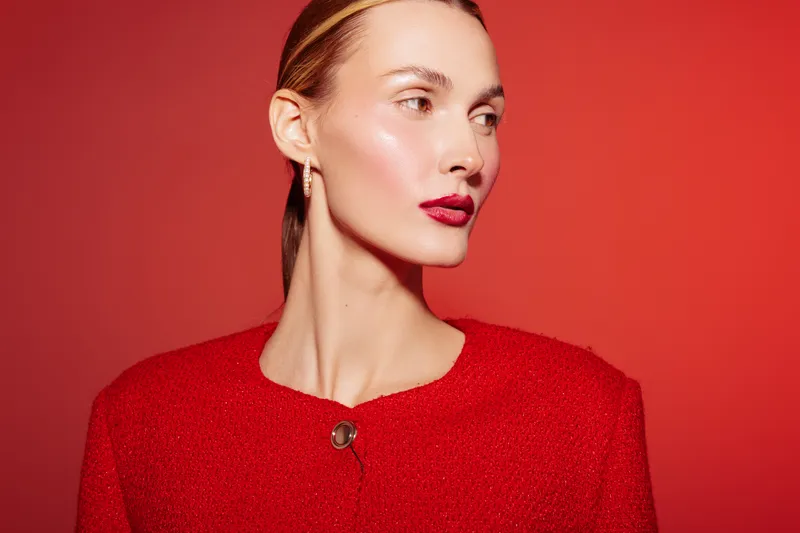 Elegant woman with glossy skin and red lipstick wears red textured jacket against red background. Fashion portrait with minimal makeup and sleek hairstyle. Concept of beauty and style.