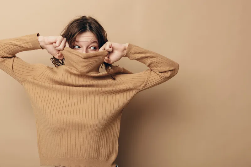 Young woman in beige knitted sweater pulls collar over face, looking sideways with playful expression. Neutral background and casual style create cozy, warm, and relaxed mood concept.