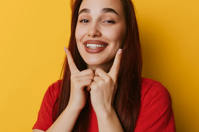 Young woman smiling and pointing fingers on yellow background for happiness, joy and positive emotion. Portrait of cheerful female with long hair in casual red shirt for lifestyle and expression.