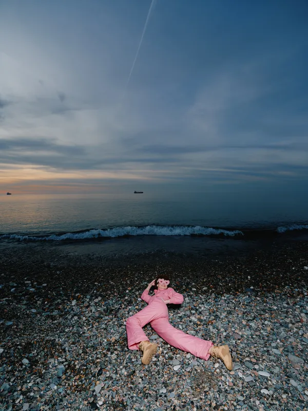 Beached scene with a person in a pink suit lying on pebbles beside the sea, during sunset. Calm horizon, gentle waves, and a distant boat create a surreal, contemplative mood on the coast.