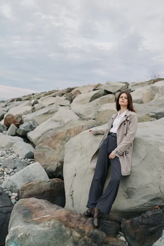 Young woman in casual outfit stands on large rocks at rocky shore under cloudy sky. Outdoors scene with natural landscape, fashion, and mood of contemplation or relaxation.
