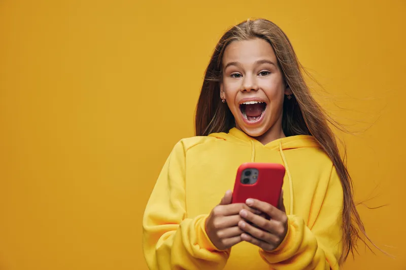 Girl with long hair wearing yellow hoodie holding red smartphone with excited expression for technology and communication on bright background with copyspace.