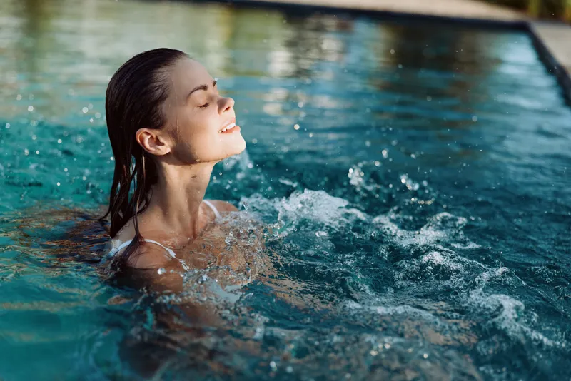Young woman with wet hair enjoying swimming pool water splash, smiling with closed eyes, lifestyle concept of relaxation and wellness outdoors under natural light.