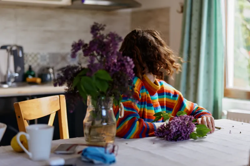 Colorful sweatered woman sits at a bright kitchen table with lilac blooms, relaxed pose, soft natural light, cozy home atmosphere, candid lifestyle moment