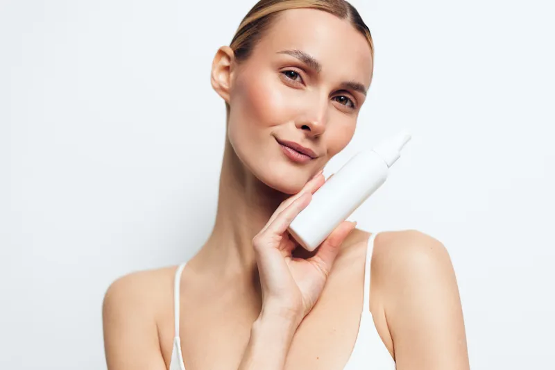 Confident woman holds a white skincare bottle, showcasing a simple beauty routine, smooth skin, soft lighting, isolated on a light background, ready to apply moisturizer