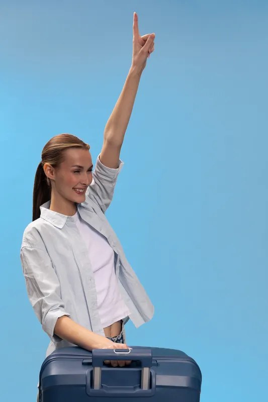 Smiling woman with suitcase raises hand happily against blue background. Concept of travel, vacation, freedom, and positive emotions with casual clothing and joyful mood.