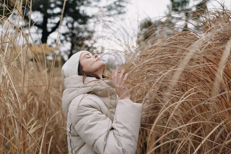 Woman in a padded coat and beanie stands among tall dried grasses, enjoying a tranquil winter moment in an outdoor natural setting, with soft light and calm atmosphere.