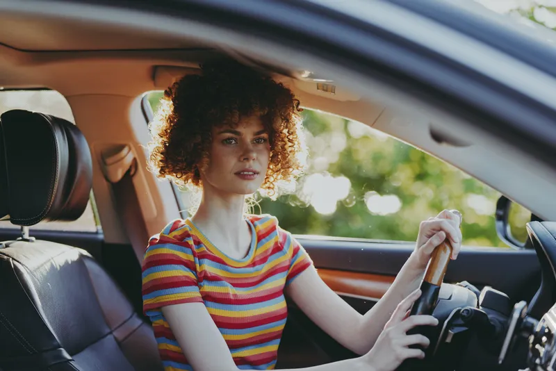 A young woman sits in a colorful striped shirt in a car, a casual, relaxed portrait captured in warm retro tones. A lifestyle moment highlighting everyday travel and self expression.