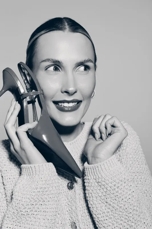 Happy white woman with light skin holding red high heel shoe near face, smiling and posing in cozy knitted sweater. Concept for fashion and lifestyle.