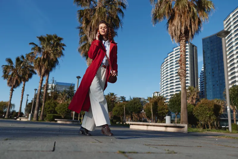 Young woman in a red coat walks confidently through a sunny city plaza, palm trees behind, wearing a white jumpsuit and black shoes, exuding bold street style.