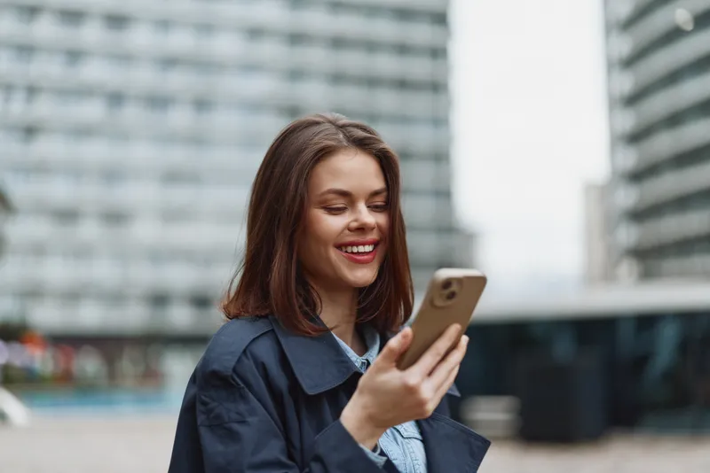Young woman in a navy jacket smiles at her smartphone in an urban setting, conveying casual connectivity and candid, focused energy.