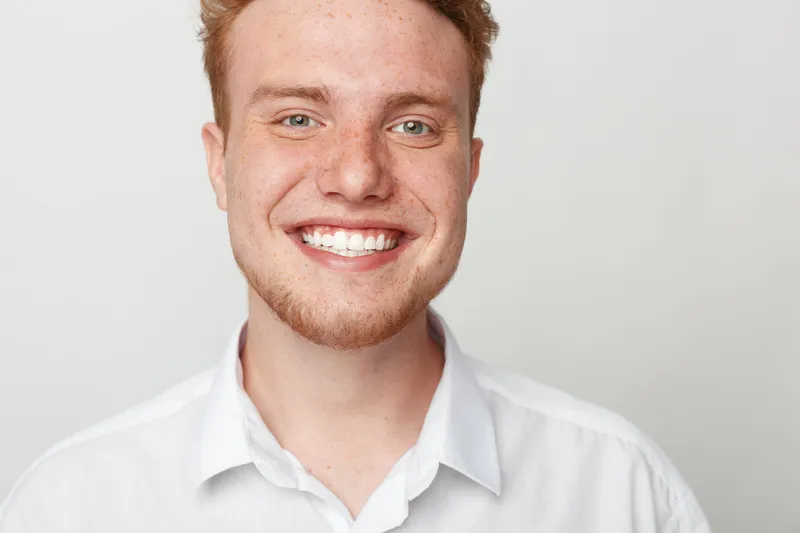 Close-up portrait of a happy young man with red hair and beard smiling broadly wearing a white shirt on a plain light background. Concept of joy, confidence, and positive emotions.