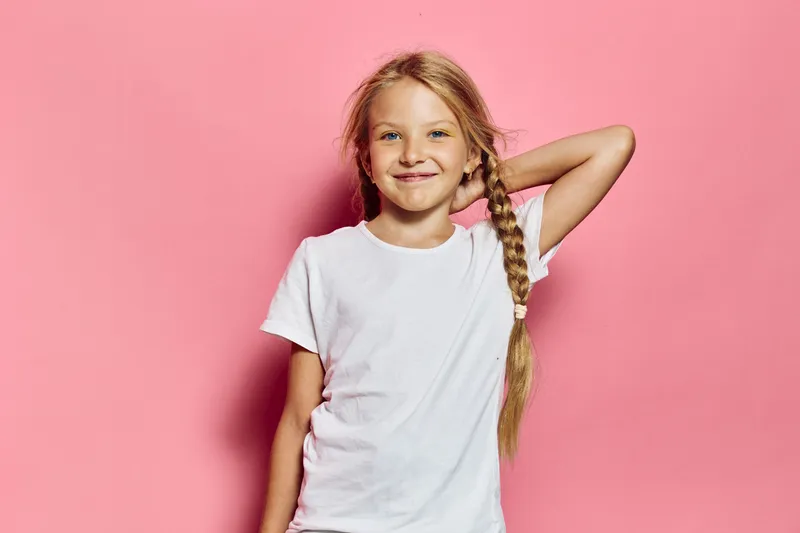 Smiling girl with long braided hair wearing a white t-shirt stands against a pink background. She poses with one hand behind her head showing confidence and happiness.