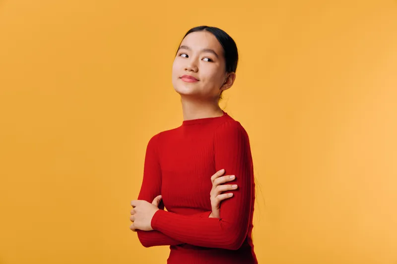 Young woman in red sweater standing with crossed arms against yellow background looking confident and thoughtful with relaxed posture and casual style in studio portrait.