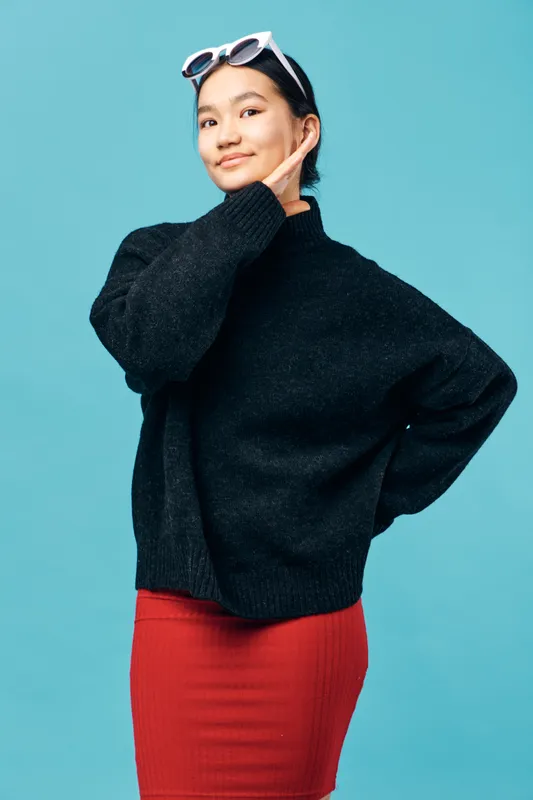 Young woman posing with hand on face wearing black sweater and red skirt against blue background. Fashionable confident model with sunglasses on head in casual style photo studio portrait.