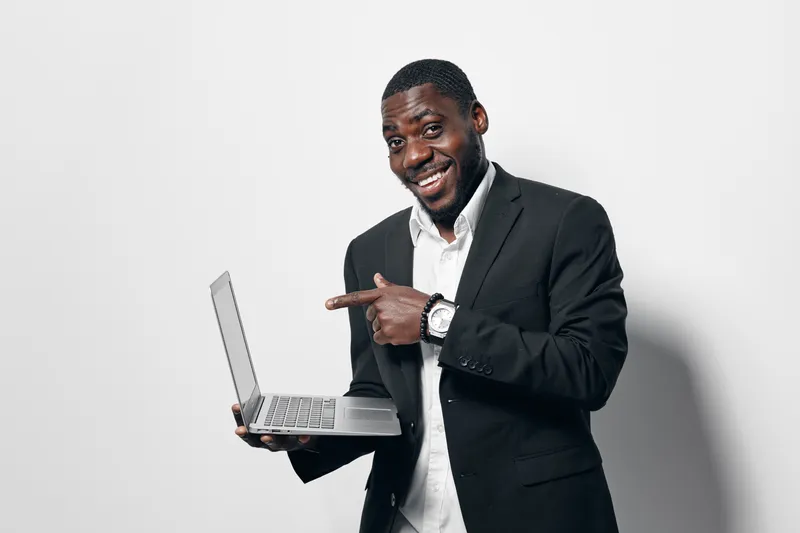 African American man with dark skin wearing black suit and white shirt smiling and pointing at laptop screen on white background. Businessman, technology and digital device for work or presentation.