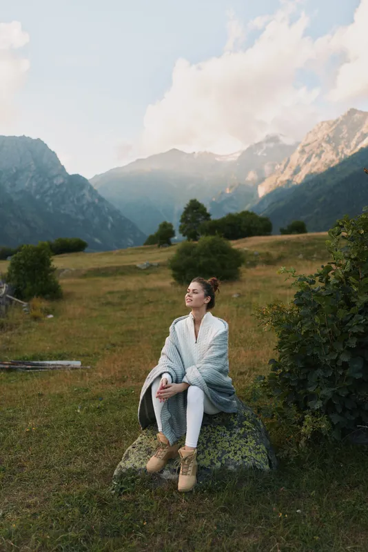 A young woman sits on a rock in a meadow with distant mountains, wearing a light cardigan and boots, looking calm and thoughtful on a sunny day.