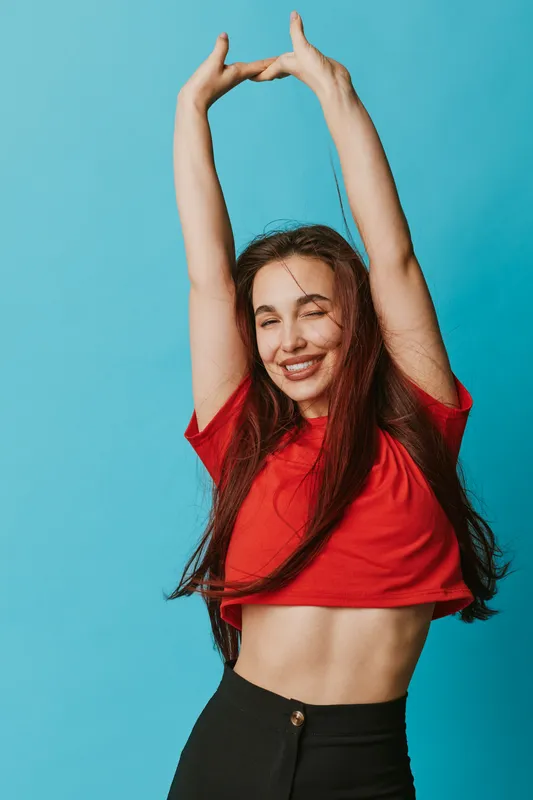 Young woman smiling with eyes closed stretching arms up wearing red top and black pants against blue background. Happy girl with long hair enjoying moment in casual style.