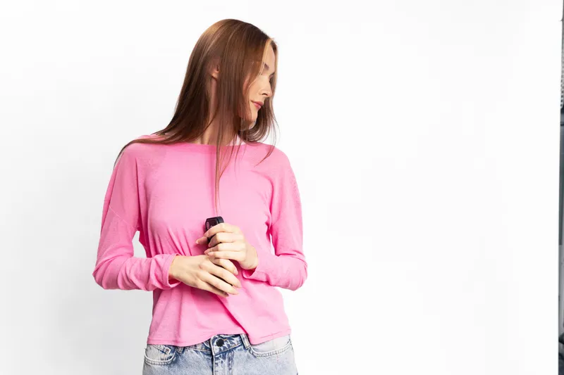 Young woman in pink sweater and jeans stands against white background holding a smartphone, looking to the side. Concept of casual style, technology use, and thoughtful mood.