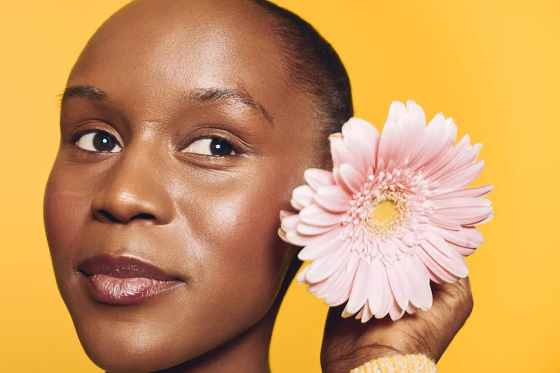 Close-up of a smiling black woman with dark skin holding a pink gerbera flower near her face on yellow background. Concept of natural beauty, skincare, and floral wellness.