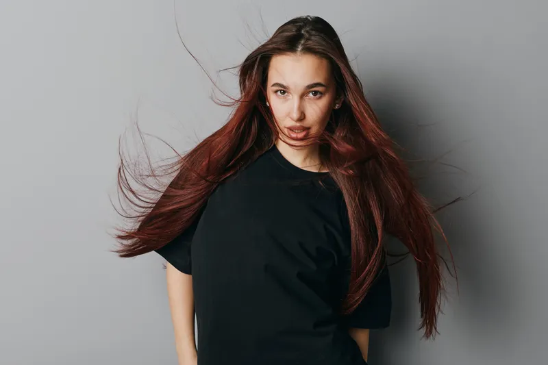 Woman with long hair blowing in wind wearing black t shirt standing against gray background with serious expression and confident posture in studio lighting.