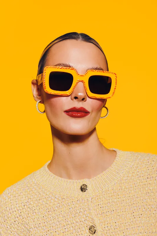 Stylish woman with slicked back hair wears large orange sunglasses and red lipstick against bright yellow background. Fashion portrait with bold colors and confident expression.