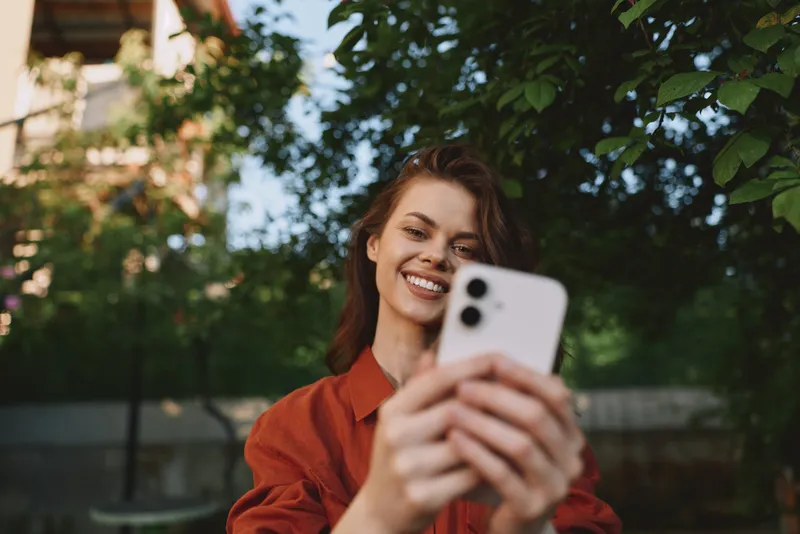 Smiling woman outdoors taking a selfie with a smartphone. Bright natural light highlights casual fashion and vibrant everyday lifestyle moments for social media