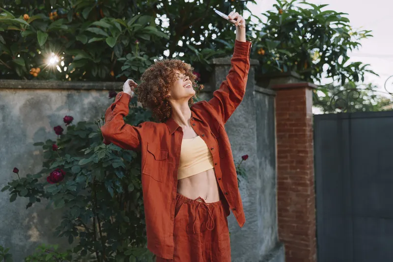 Joyful woman lifestyle outdoor in red outfit raises hands toward the sky, a sunny garden portrait with curly hair, confident pose and energetic expression and joy