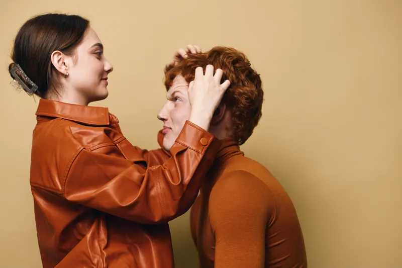 Woman adjusting hair of man with curly hair in brown sweaters against beige background indoors, focusing on care and close relationship between two people.