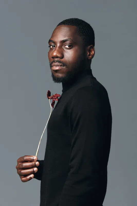 African man with dark skin in creative conceptual visual style holding dried flower wearing black turtleneck against gray background studio portrait with serious expression.
