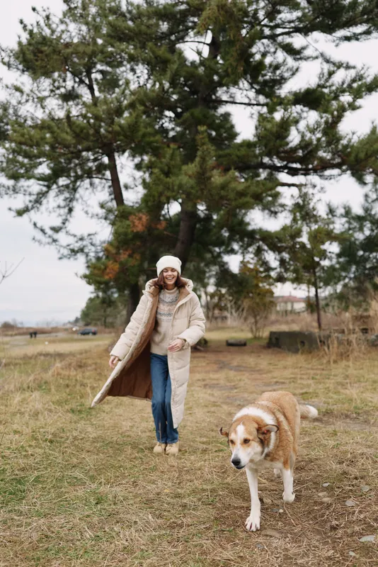 Woman walking a dog in a park on a cool day bundled in a warm coat and hat leash in hand open field and trees behind for a calm outdoor scene