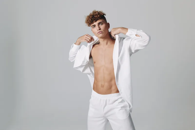 Young man with curly hair posing in white pants and open white shirt showing toned abs against gray background in studio for fashion and fitness concept.