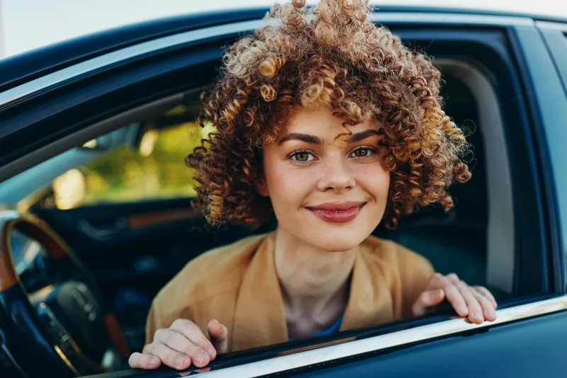 Bright portrait of a woman leaning from a car window, smiling at the camera, outdoors in warm retro tones, casual chic outfit, soft breeze, natural greenery background