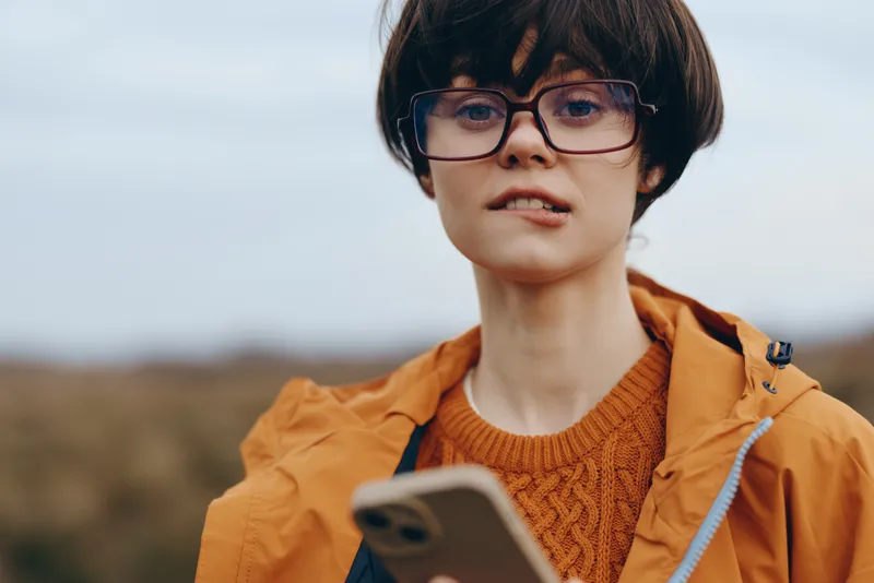 Woman with glasses outdoors holding smartphone, portrait in countryside field, wearing orange jacket and casual knit sweater, natural daylight lifestyle moment, soft wind, attentive expression