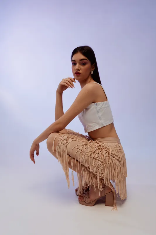 Young woman in a white crop top and beige fringed skirt poses crouching on a plain background. Fashion concept focused on beauty, style, and modern trends.
