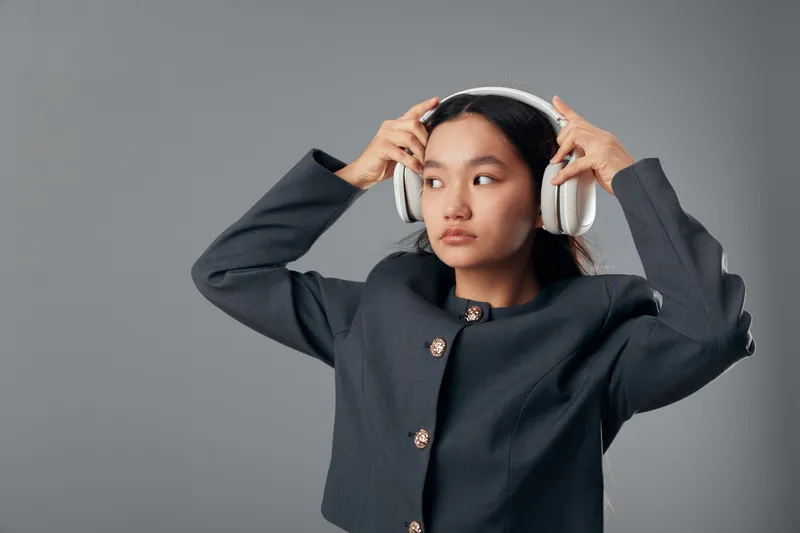 Young woman wearing headphones adjusting them on head looking away with thoughtful expression in studio. Portrait, technology, music, fashion, casual, lifestyle and modern concept.