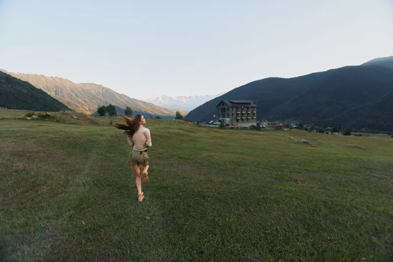 Young woman runs across a wide countryside field toward a distant building, mountains rising behind, conveying an active lifestyle, outdoors joy, and freedom.