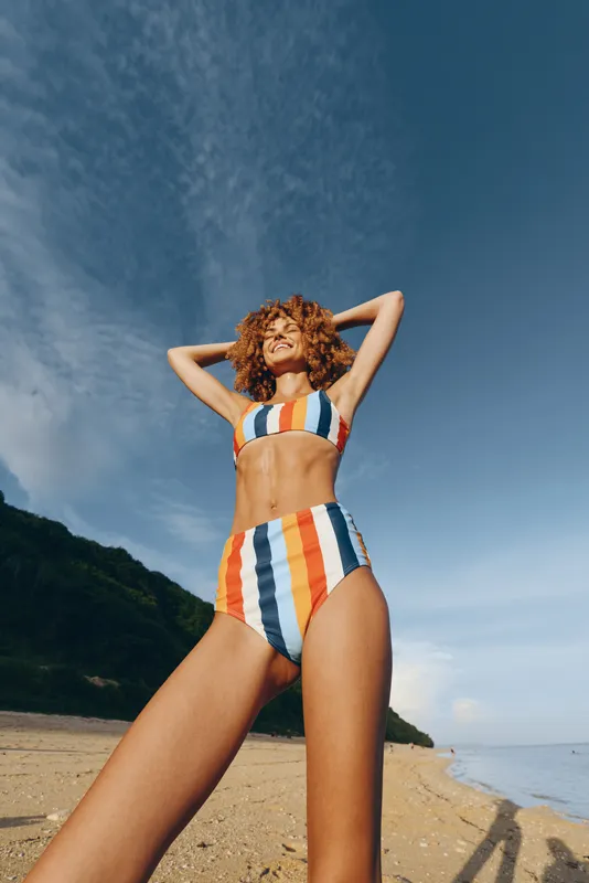 Happy young woman in colorful striped bikini stands on sandy beach with hands behind head, enjoys sunny day with blue sky and green hill in background.