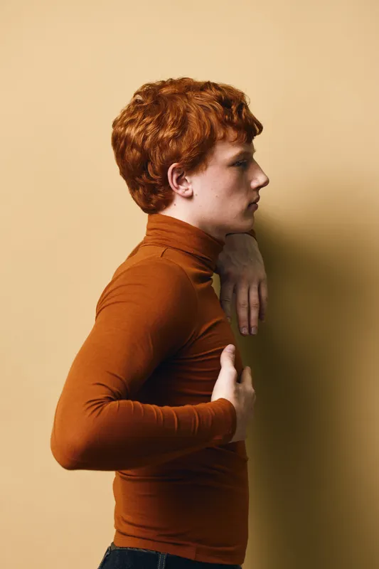 Young man with red hair wearing brown turtleneck posing sideways against beige wall, showing profile and thoughtful expression in stylish casual outfit indoors.