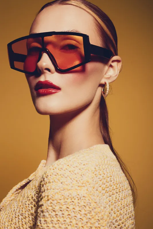 Stylish woman with slicked back hair wears oversized tinted sunglasses and hoop earrings. She poses confidently against a warm yellow background in a textured knit top. Fashion concept.