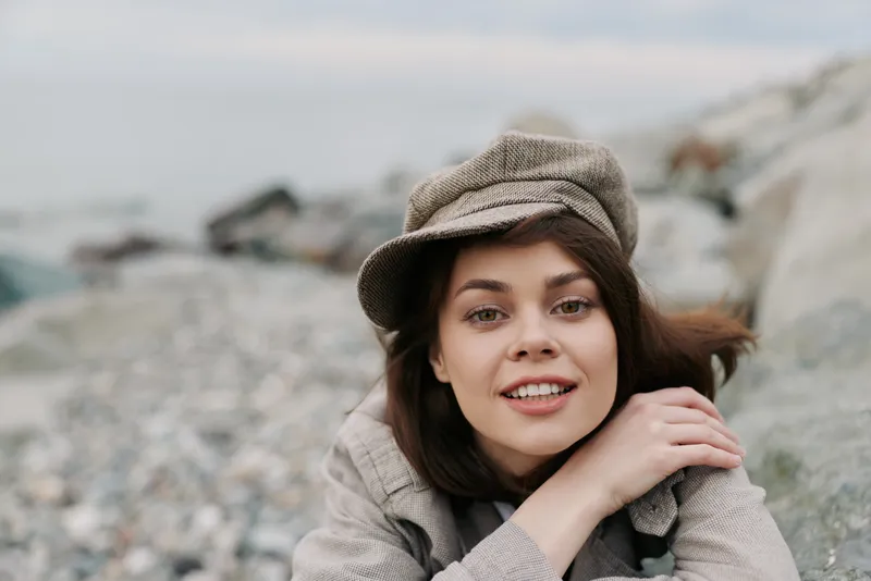 Young woman with brown hair wearing a beige hat and coat smiles gently while lying on rocky beach. Natural light highlights her clear skin and relaxed expression outdoors.