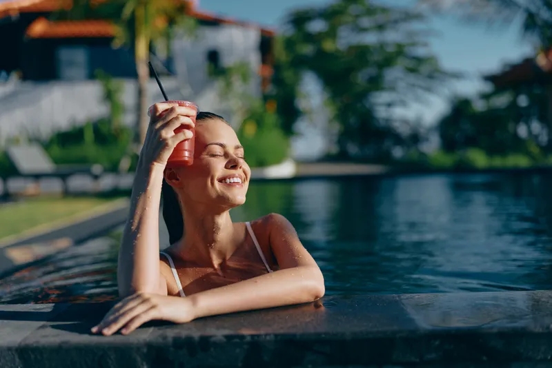 Cheerful woman in a sunlit outdoor pool, smiling with eyes closed, relaxed pose, clear water and lush surroundings, warm breeze, casual swimwear, tranquil moment, vibrant colors