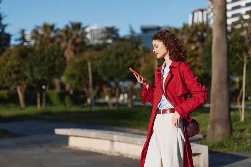 Stylish woman in a red trench coat checks her tablet in a sunny park, wearing white pants and a brown crossbody bag, exuding confidence and casual elegance.
