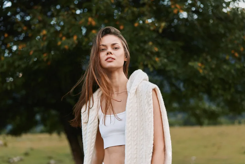 A young woman in a white crop top with a knitted cardigan draped over her shoulders stands in a sunlit field, exuding casual confidence, with hair moving softly in the breeze.