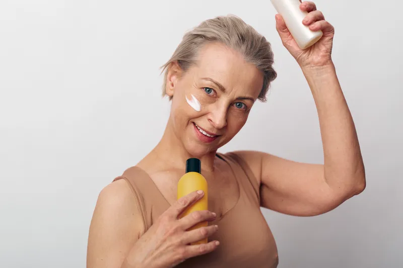 Smiling mature woman with natural skin applies cream on face holding cosmetic bottles in both hands against plain background. Concept for skincare routine, beauty care, and self-care habits.