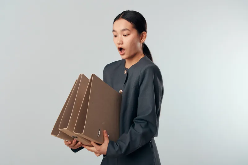 Young woman holding multiple cardboard boxes with surprised expression on face. Delivery, packaging and shipping concept with female person in casual dark clothing against light background.