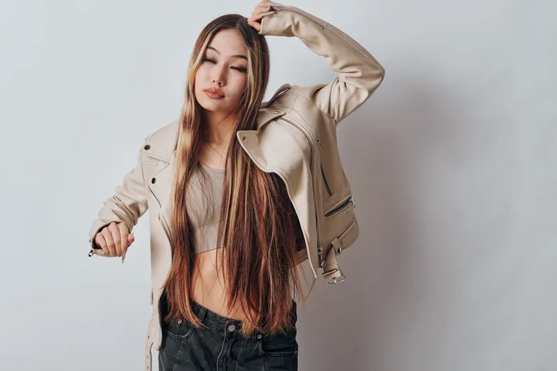 Young woman with long hair wearing beige jacket and crop top posing with closed eyes in casual style on light background in studio portrait