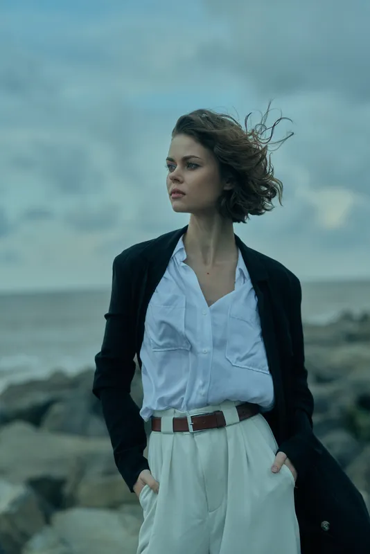 Windy coastal scene with a woman in a blazer and white shirt, hands in pockets, gazing toward the horizon, projecting professional style and calm confidence.